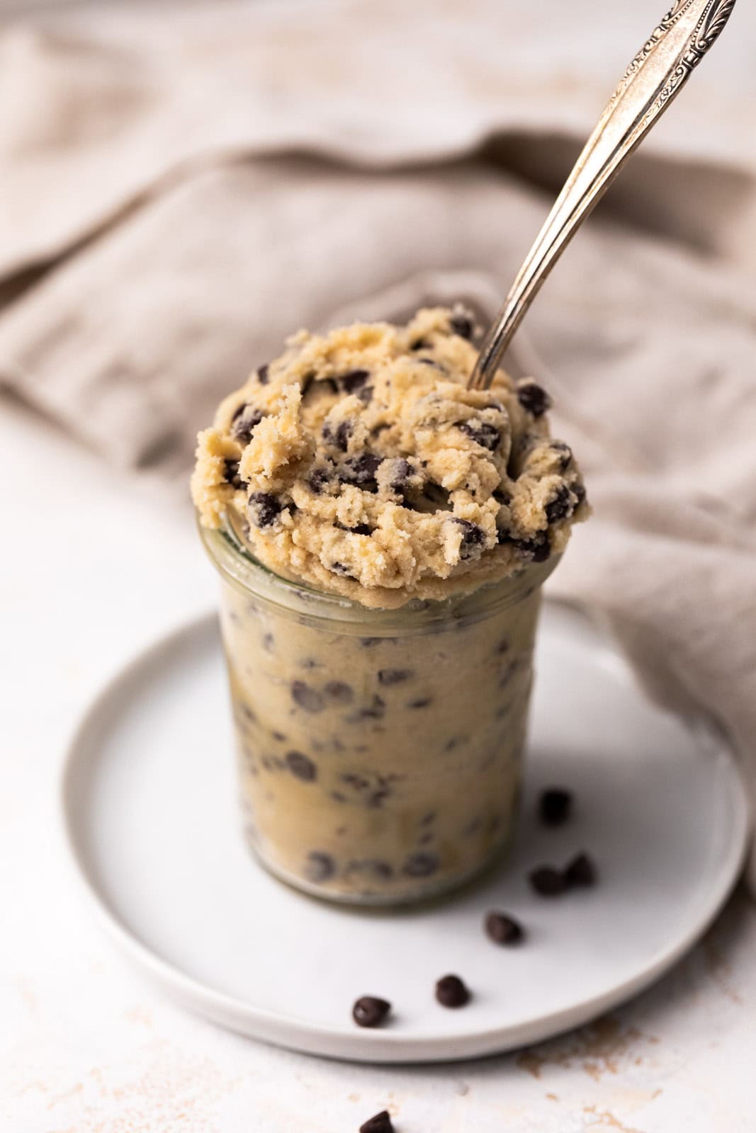 Close up of edible chocolate chip cookie dough in a glass jar on a white plate with a spoon coming out of the cookie dough.