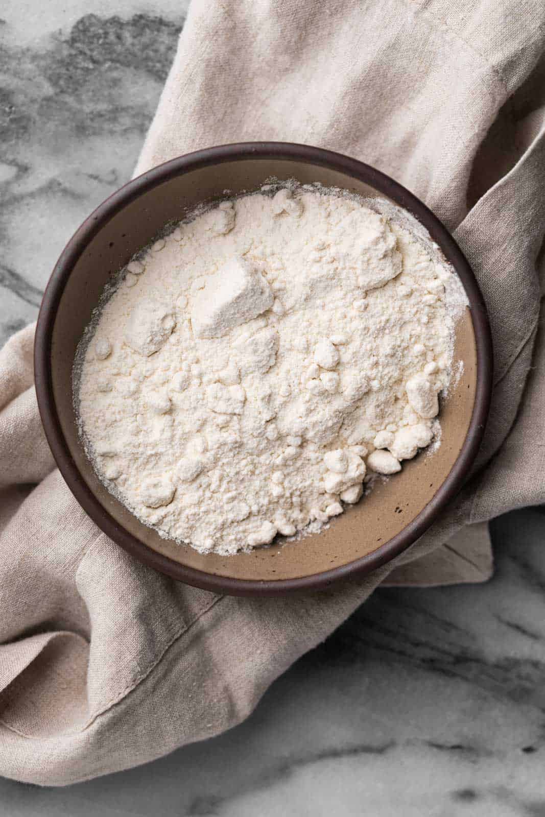 Heat-treated flour in a bowl bowl set on a linen napkin on a marble flat surface.