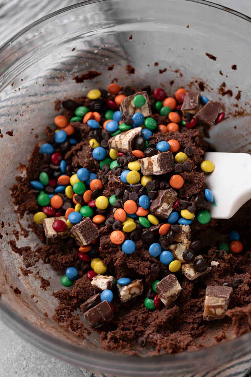 Edible chocolate cookie dough with mini M&Ms and chopped candy in a glass mixing bowl with a white spatula.