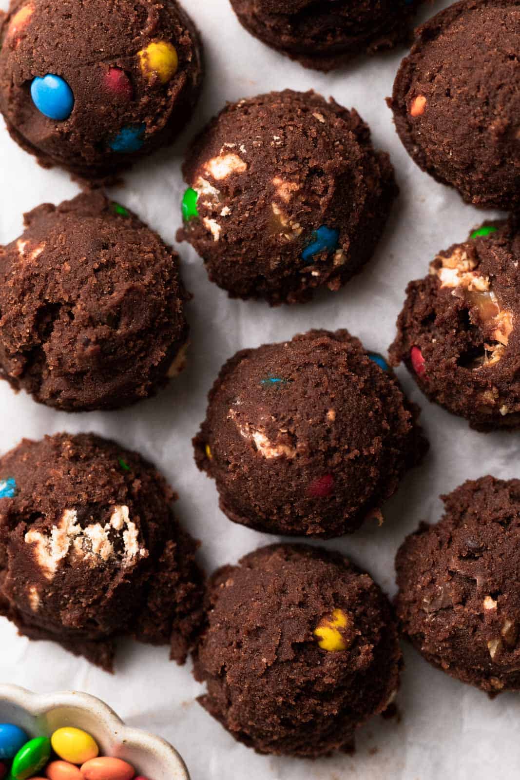 Scoops of edible chocolate cookie dough mixed with candy on a parchment sheet.