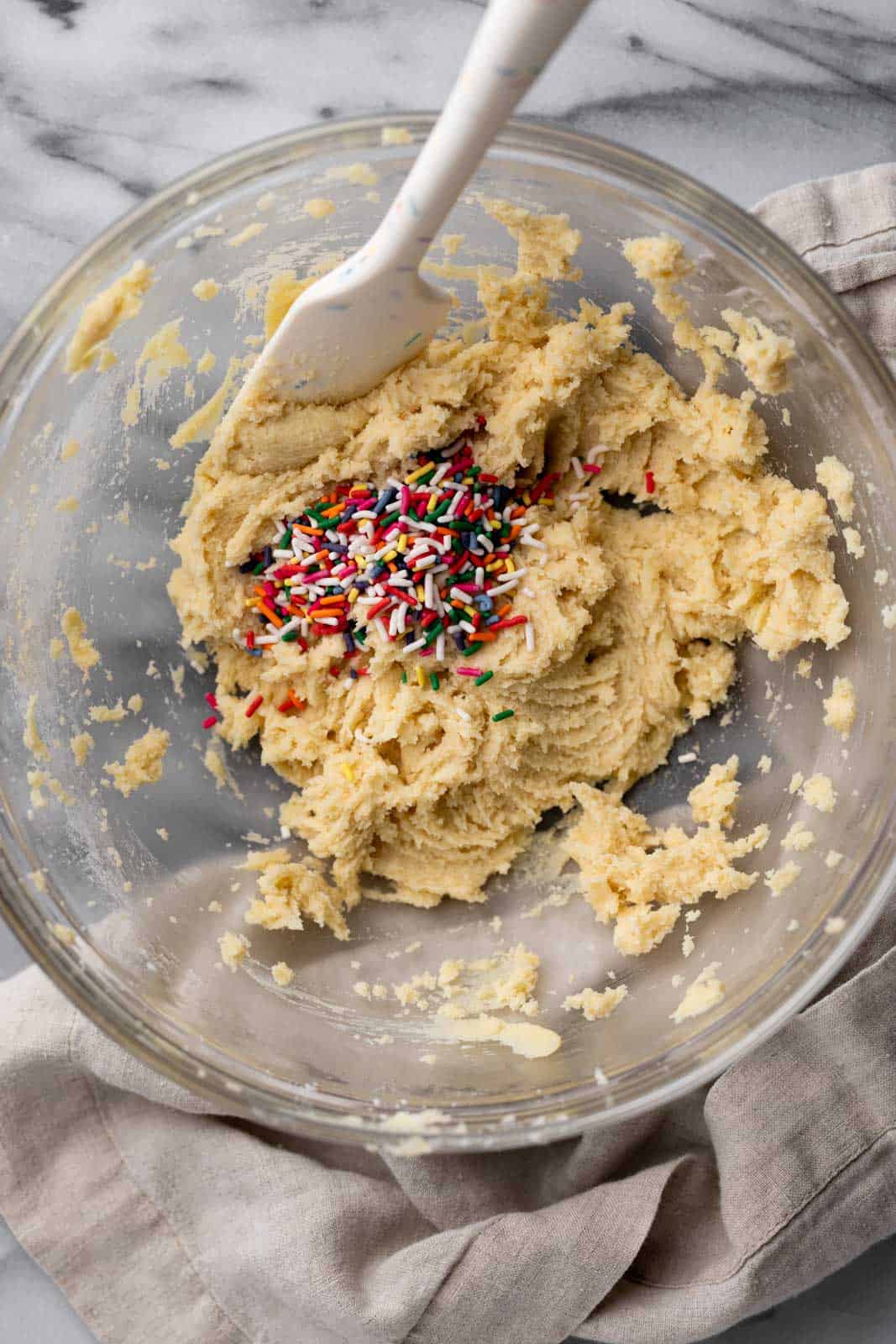 Edible sugar cookie dough in a glass mixing bowl with a white rubber spatula and sprinkles on top.