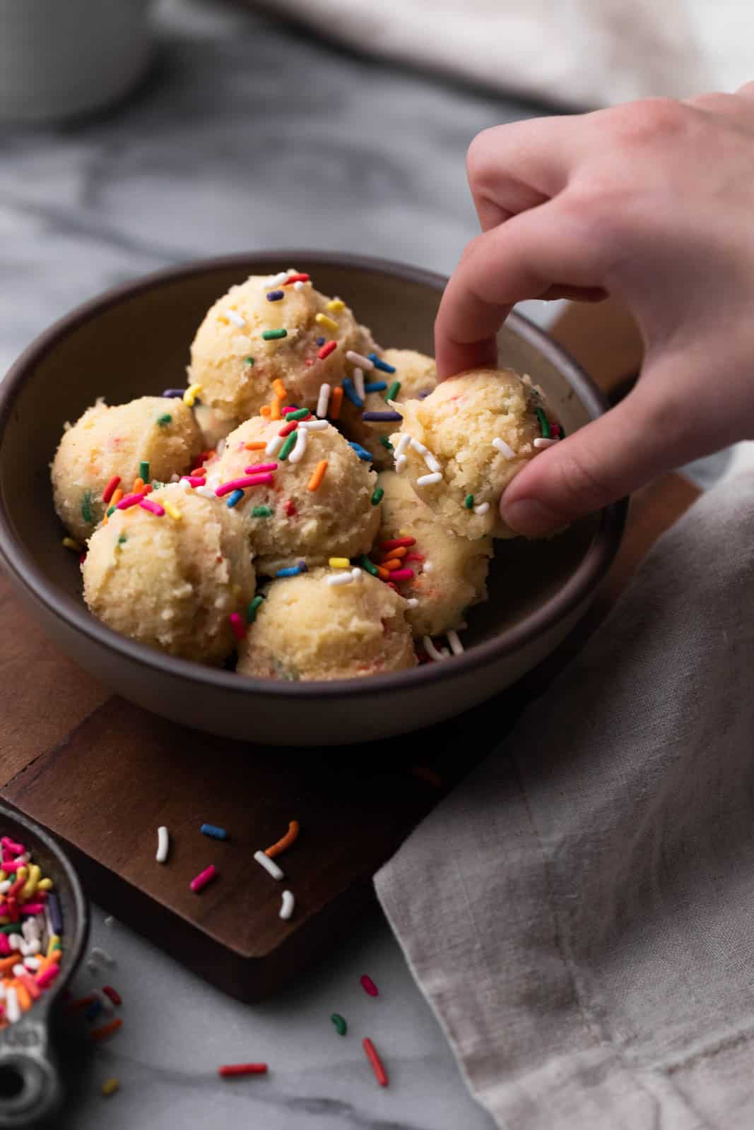 Brown bowl with scoops of edible cookie dough inside and sprinkles on top with a hand grabbing a cookie dough ball.
