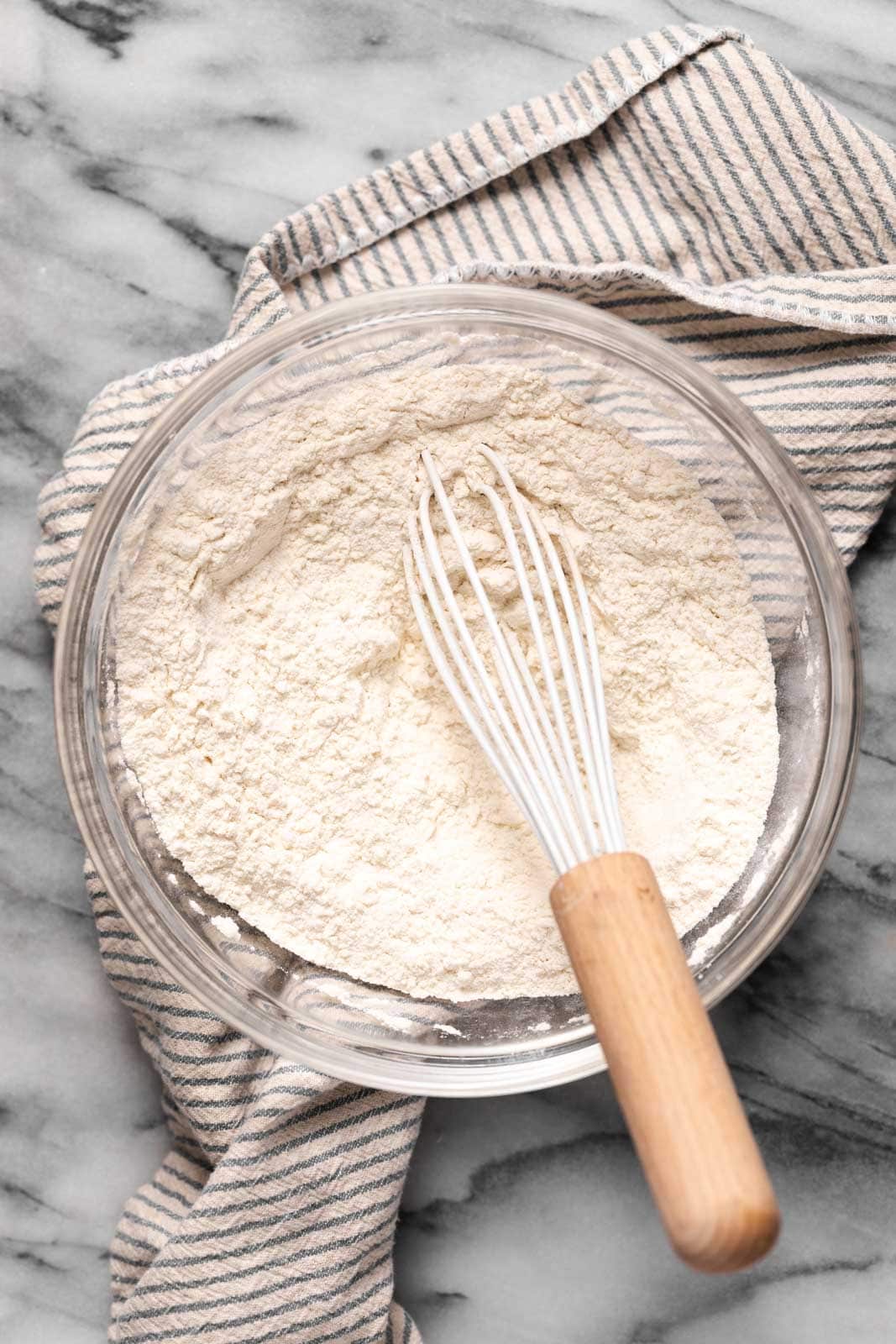 Whisked dry ingredients in a glass mixing bowl with a wooden handled whisk all set on top of a striped linen on a marble surface.