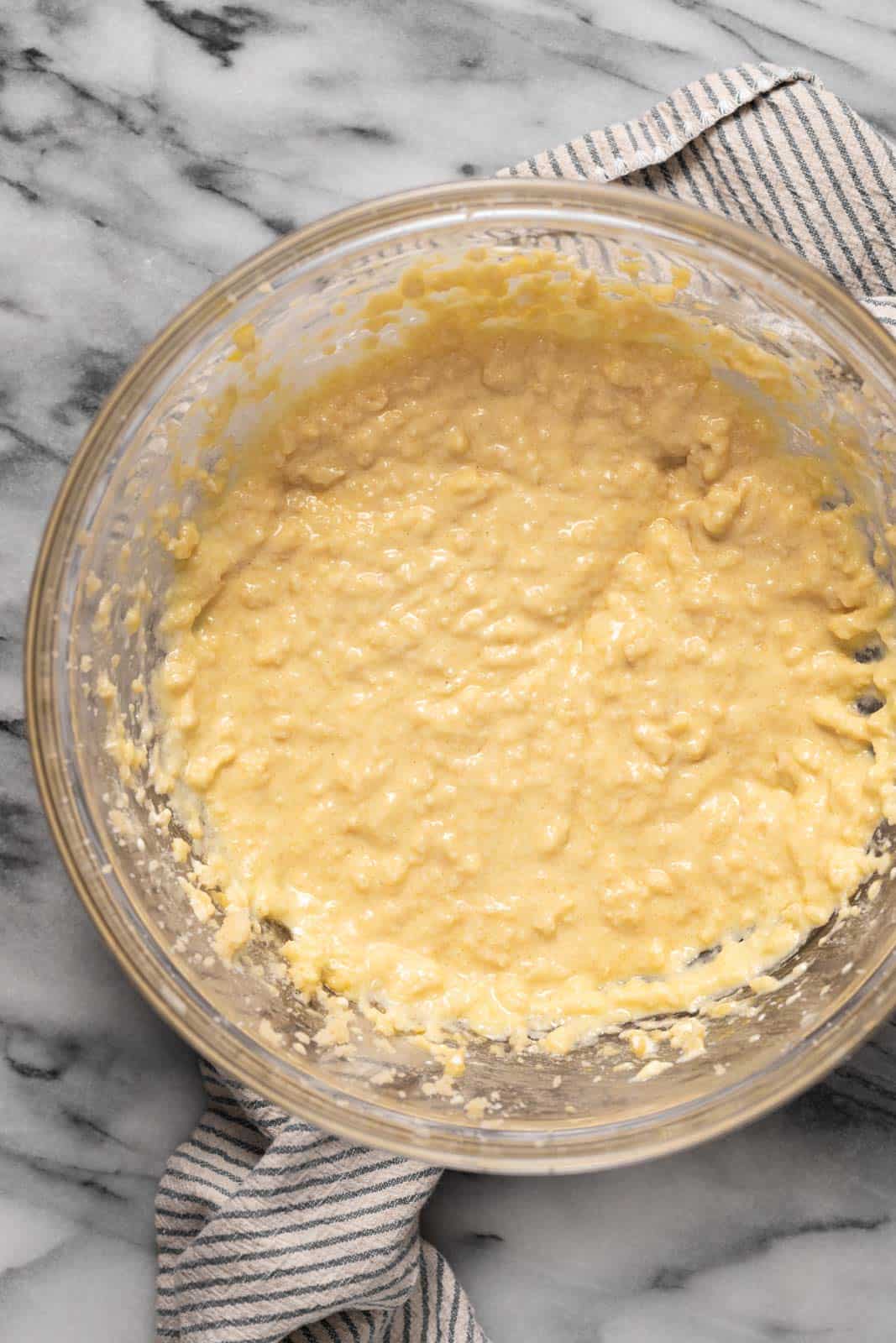 Creamed butter and sugar mixed with eggs, vanilla and sour cream in a glass mixing bowl set on top of a striped linen on a marble surface.