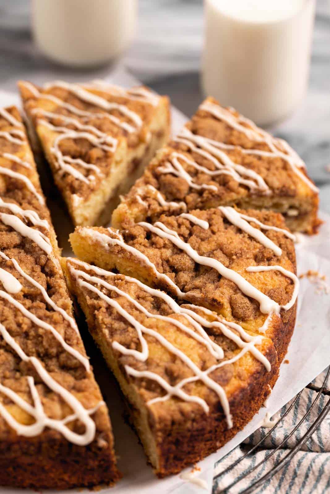 Sour cream coffee cake set drizzled with vanilla glaze set on parchment paper on a round wire rack and half sliced into triangles.