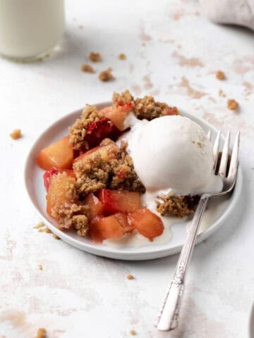 Baked strawberry apple crisp in a white round plate with a fork and scoop of vanilla ice cream with a glass of milk.
