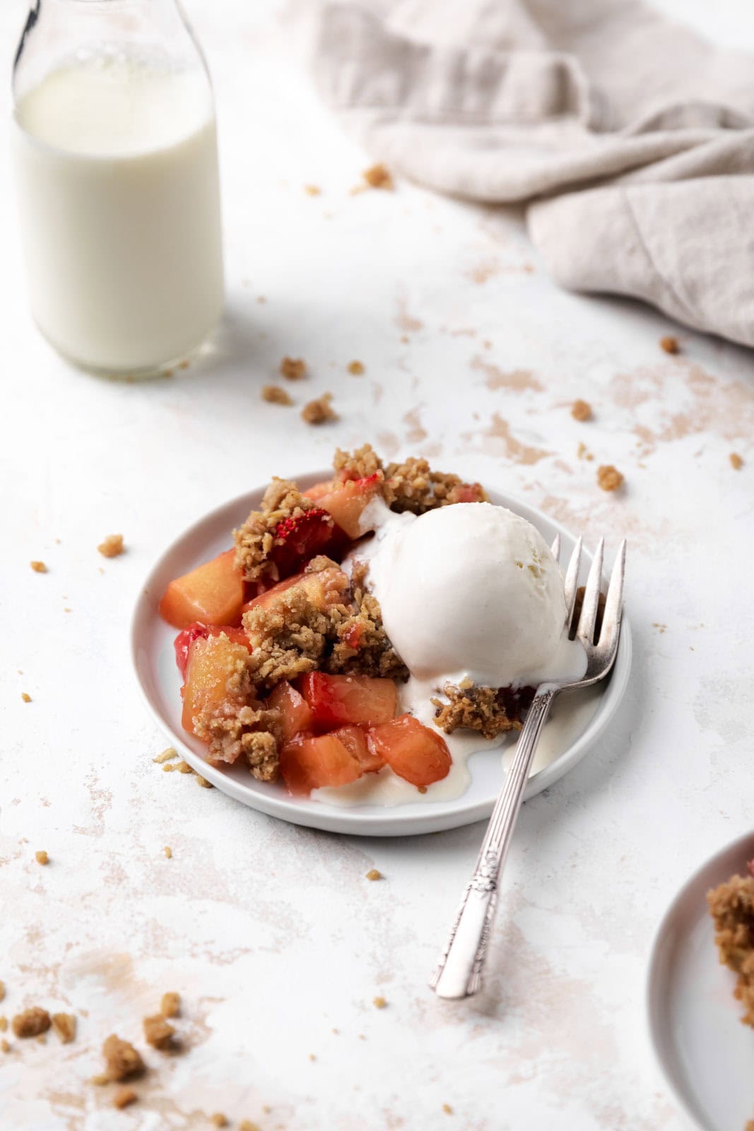 Baked strawberry apple crisp in a white round plate with a fork and scoop of vanilla ice cream with a glass of milk.