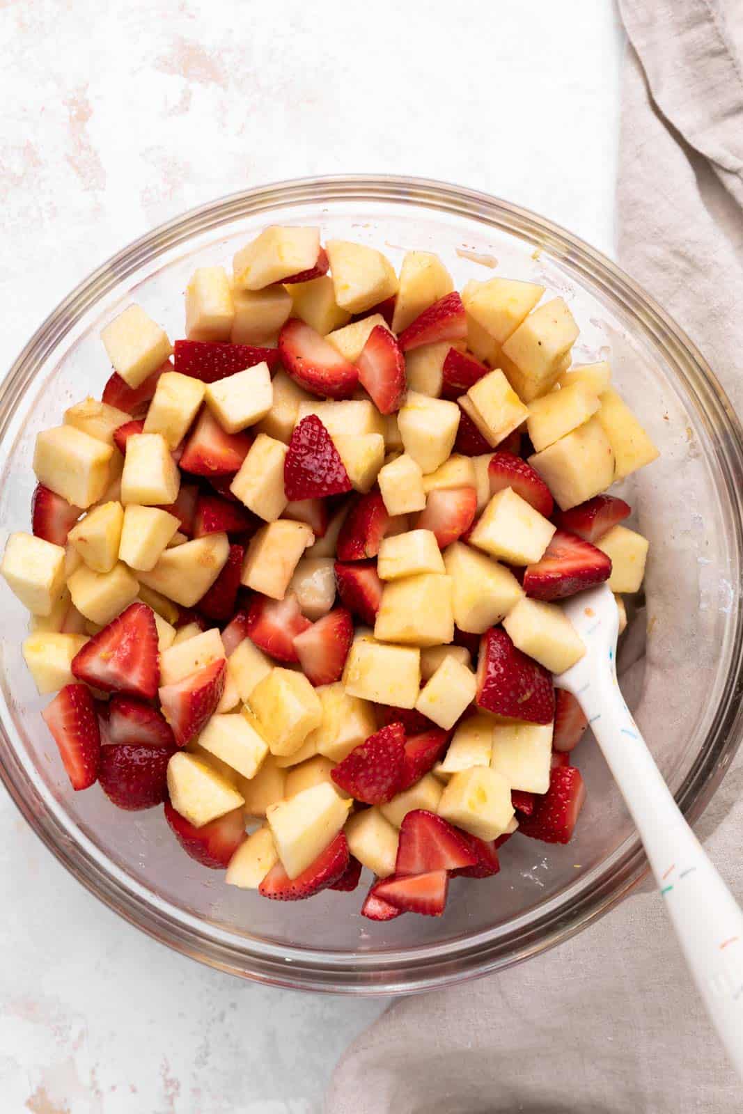 Chopped apples and quartered strawberries tossed with sugar, lemon zest and cornstarch in a glass mixing bowl set on a flat surface.