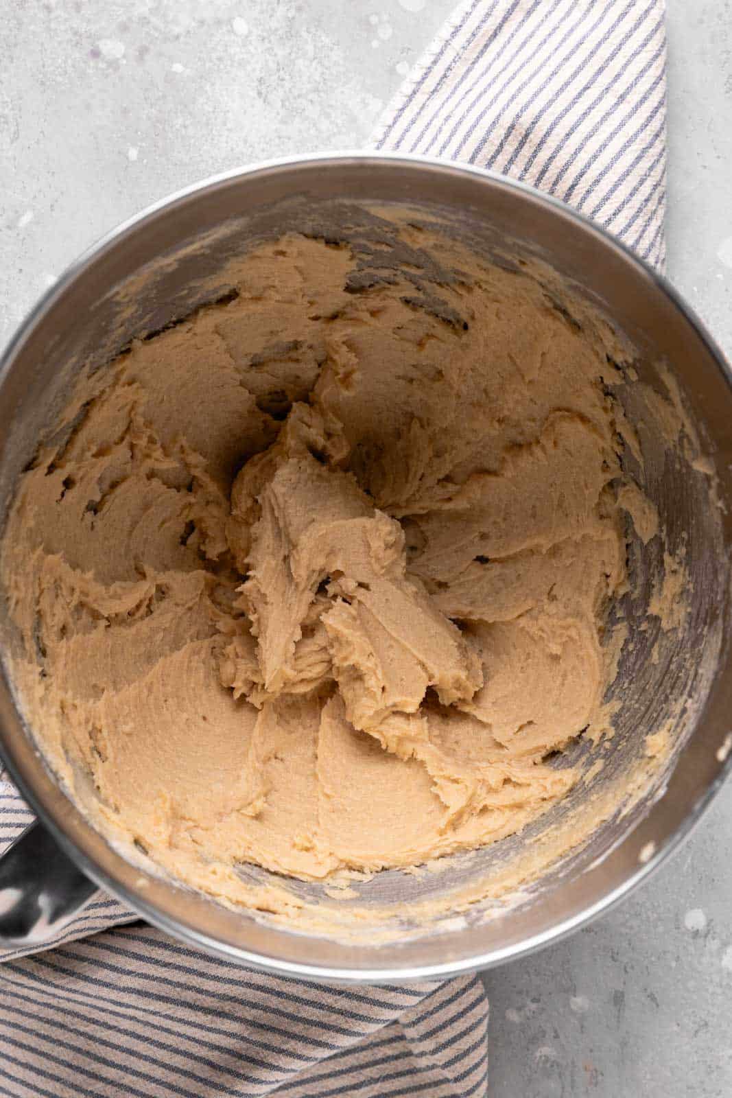 Creamed butter and brown sugar in a stand mixer metal bowl set on a flat surface with a striped linen.