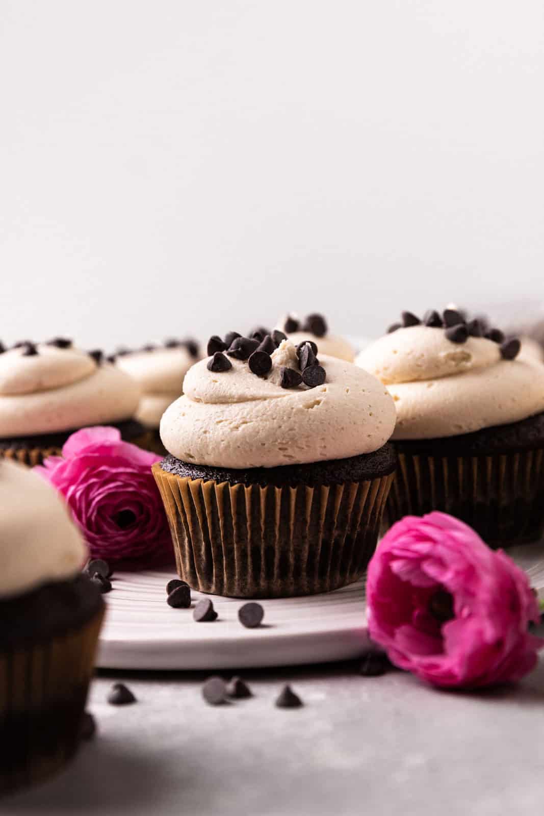 Close up of chocolate cupcakes topped with cookie dough buttercream and sprinkles of mini chocolate chips arranged on a round white dish with pink flowers around.