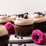 Close up of chocolate cupcakes topped with cookie dough buttercream and sprinkles of mini chocolate chips arranged on a round white dish with pink flowers around.