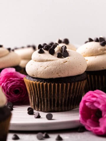 Close up of chocolate cupcakes topped with cookie dough buttercream and sprinkles of mini chocolate chips arranged on a round white dish with pink flowers around.