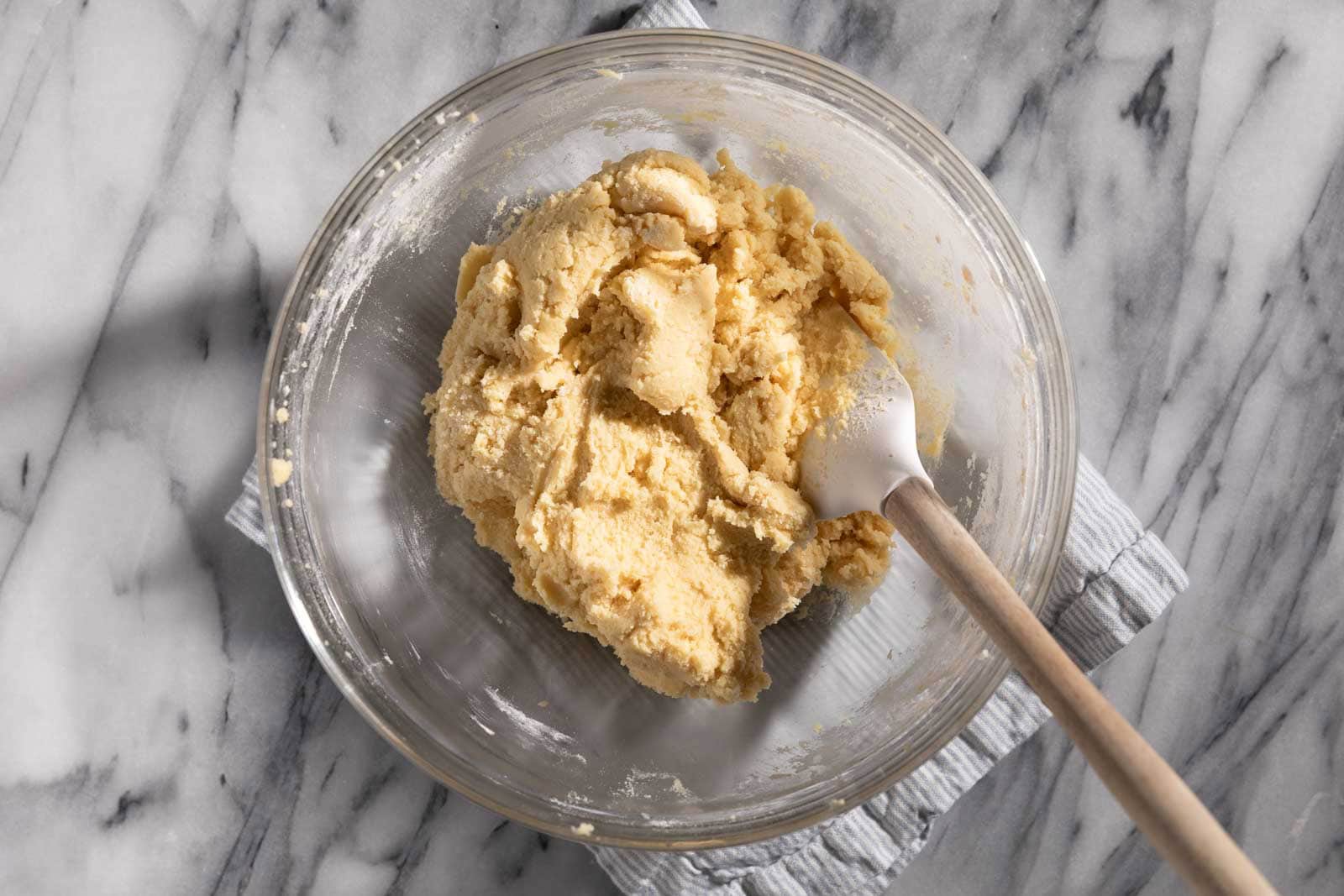 Mixed sugar cookie dough in a glass mixing bowl with a rubber spatula all set on a marble slab.