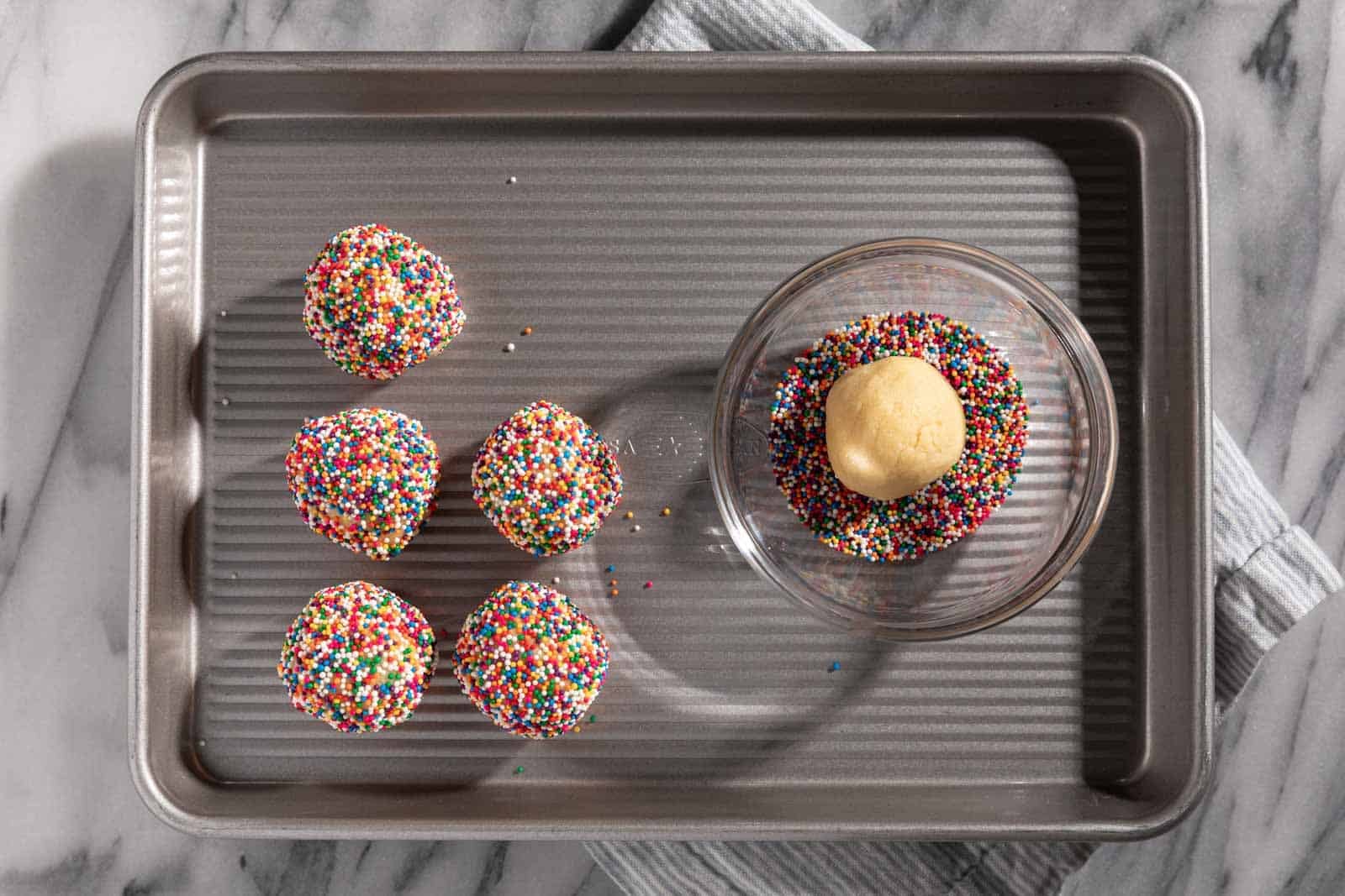 Baking sheet tray with sprinkle sugar cookie dough balls rolled in sprinkle nonpareils with a bowl of sprinkles and dough ball in it.